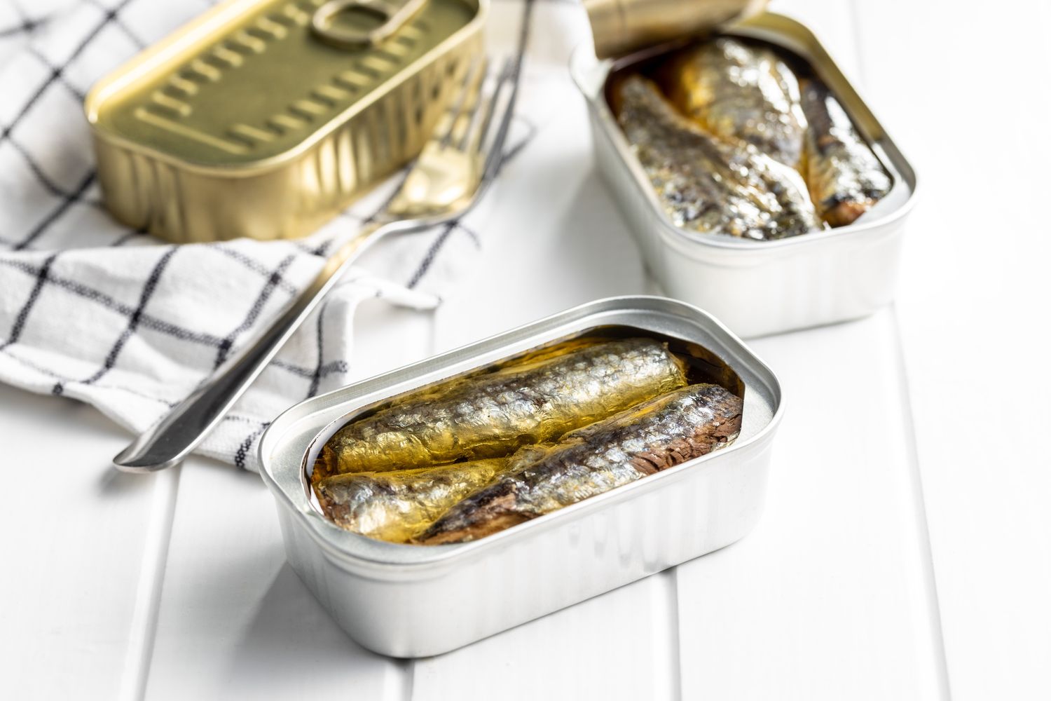 Three open cans of sardines in oil on a white wooden surface with a fork.