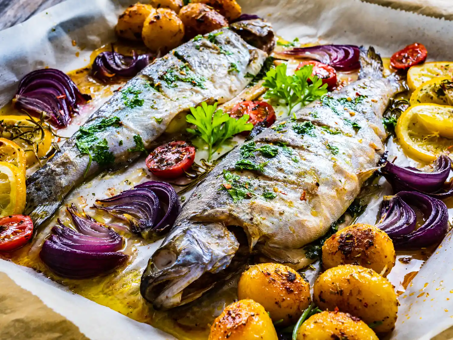 Baked whole fish with roasted potatoes, red onions, and cherry tomatoes on parchment paper.