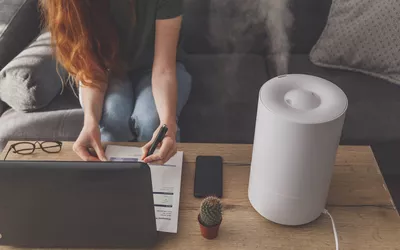 A woman working at a laptop in a home office with a humidifier emitting steam in the background.