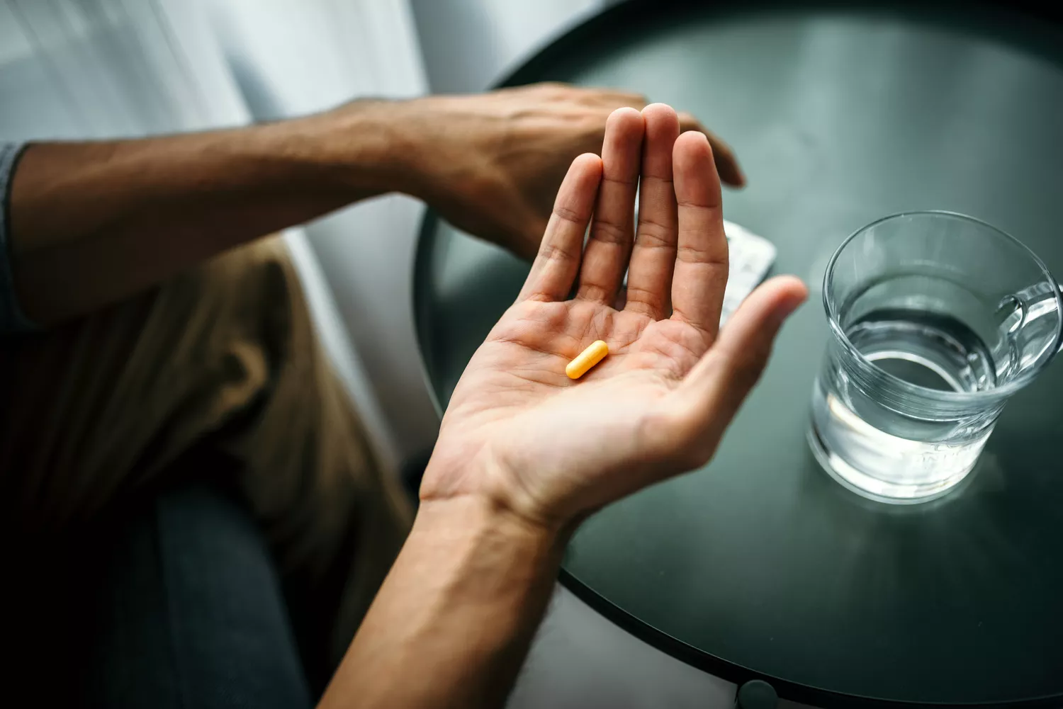 A close-up shot of a hand holding a single pill over a table with a glass of water, representing medical or supplemental factors.