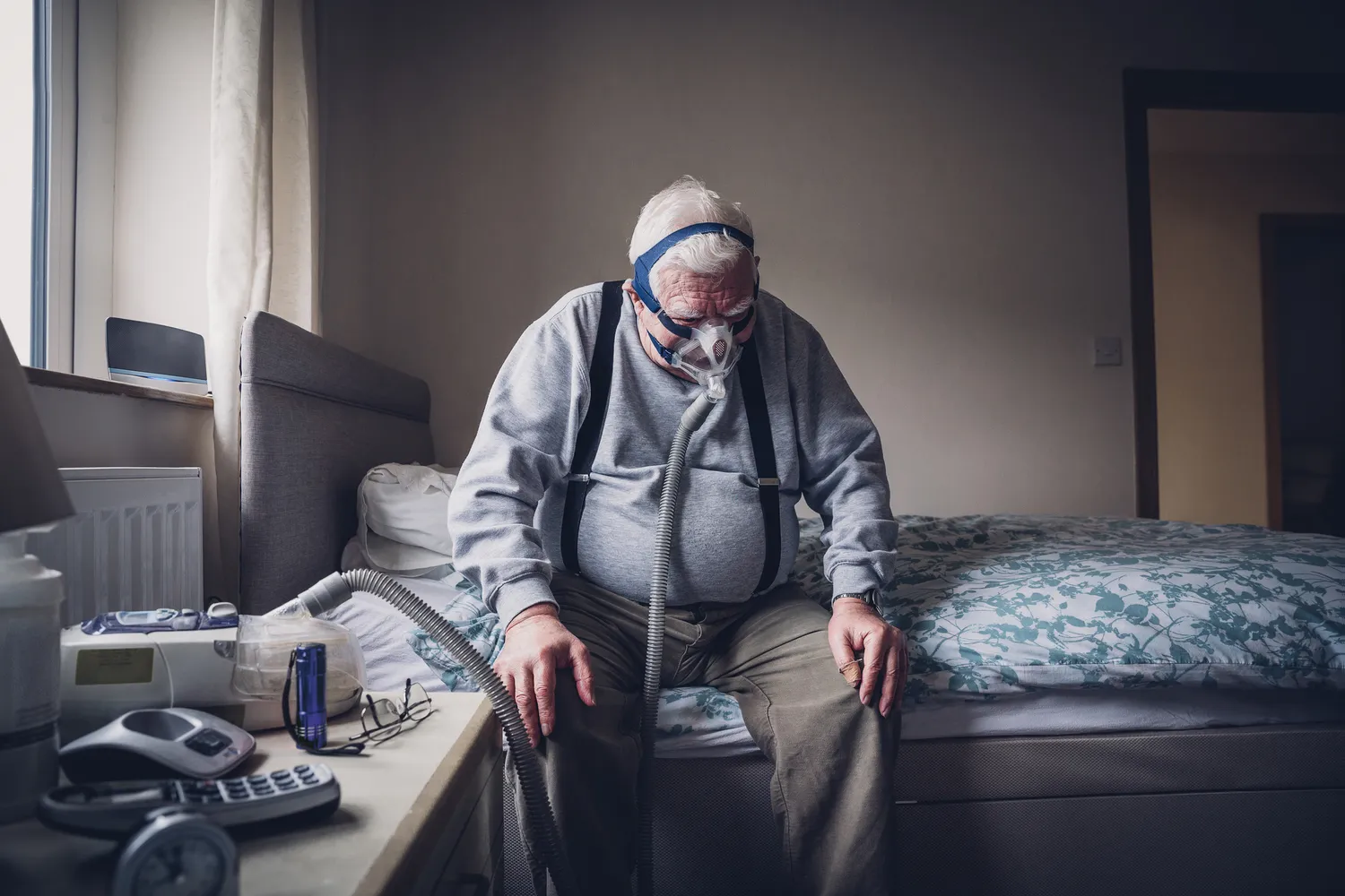 A senior man with a CPAP machine and mask in a dimly lit bedroom setting.