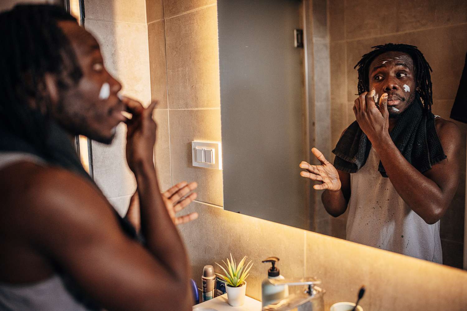 A man with dreadlocks performing a morning skincare routine in a well-lit bathroom.