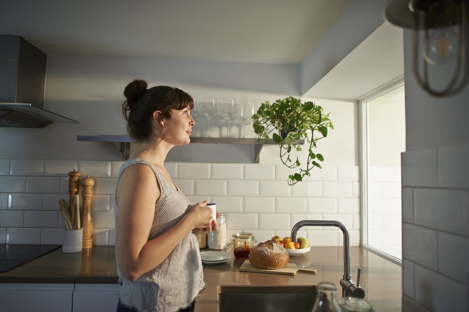 A woman stands in a sunlit kitchen with a coffee mug and a breakfast spread on the counter.