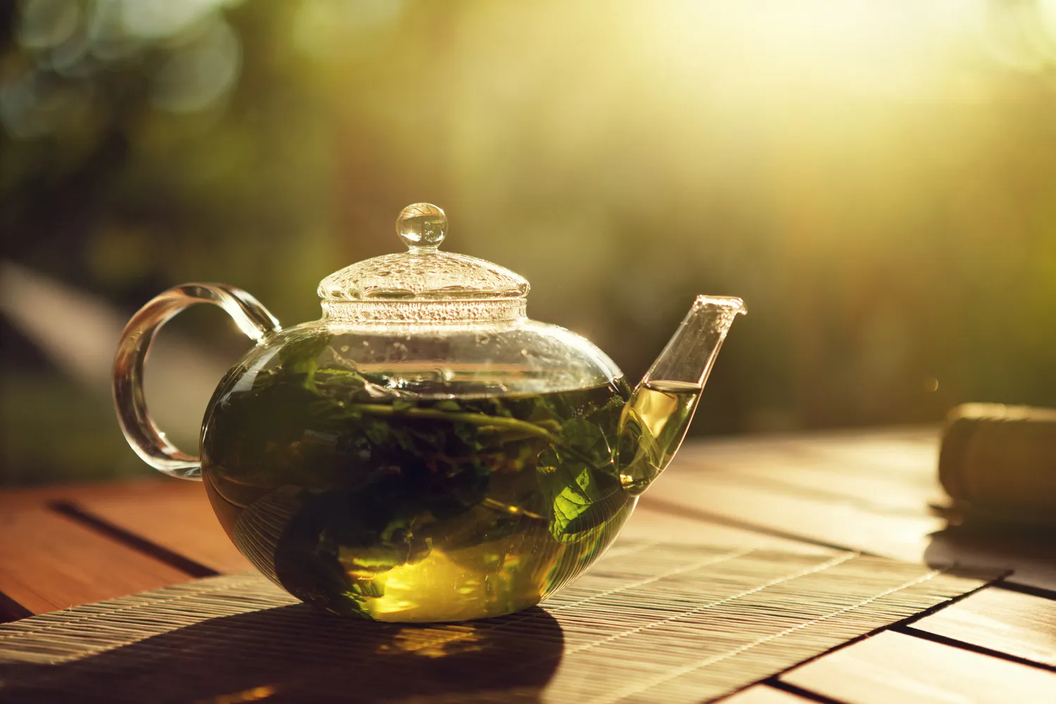 A clear teapot of green tea steeping in warm sunlight on a bamboo mat.