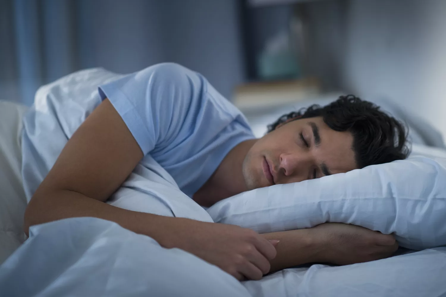 An adult man sitting on the edge of his bed looking concerned during the night.