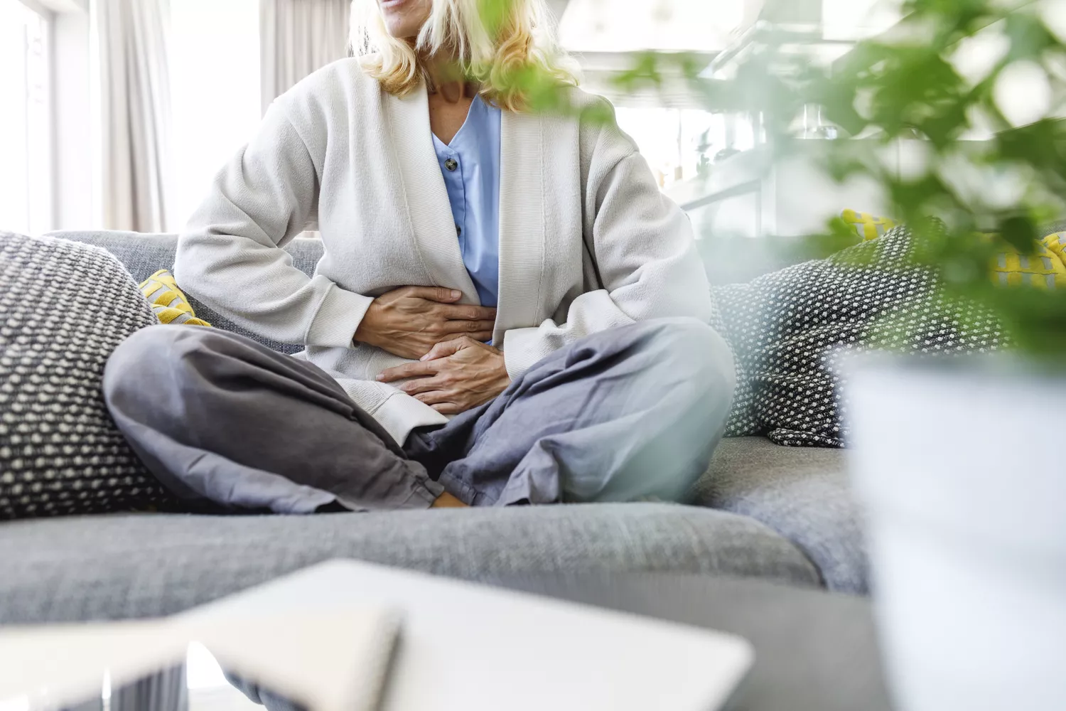 A mature woman sitting on a sofa holding her stomach in discomfort.
