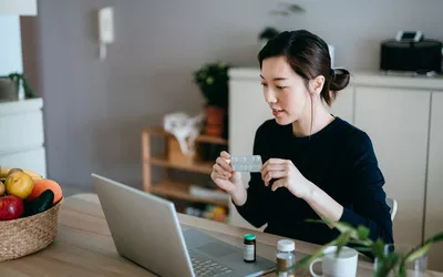 A woman reviewing medication bottles during a telehealth video call on her laptop.