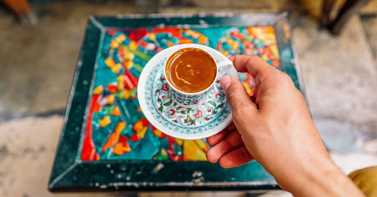 A hand holds a traditional ornate cup of espresso on a colorful mosaic tray.