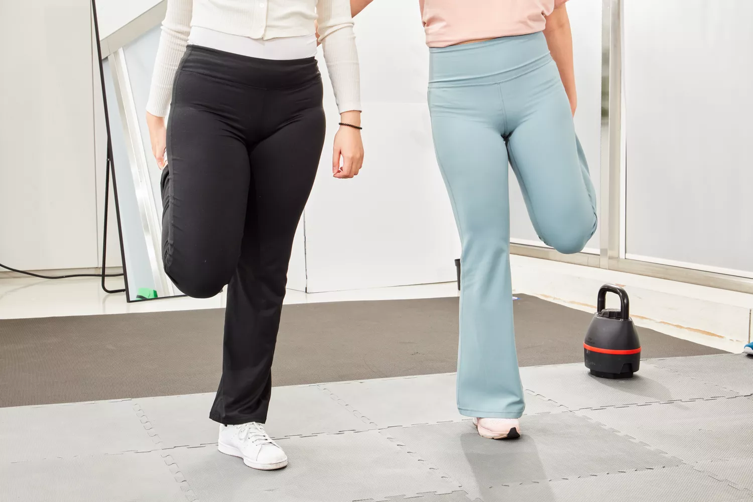 Two women performing standing quad stretches in a bright gym setting wearing athletic leggings.