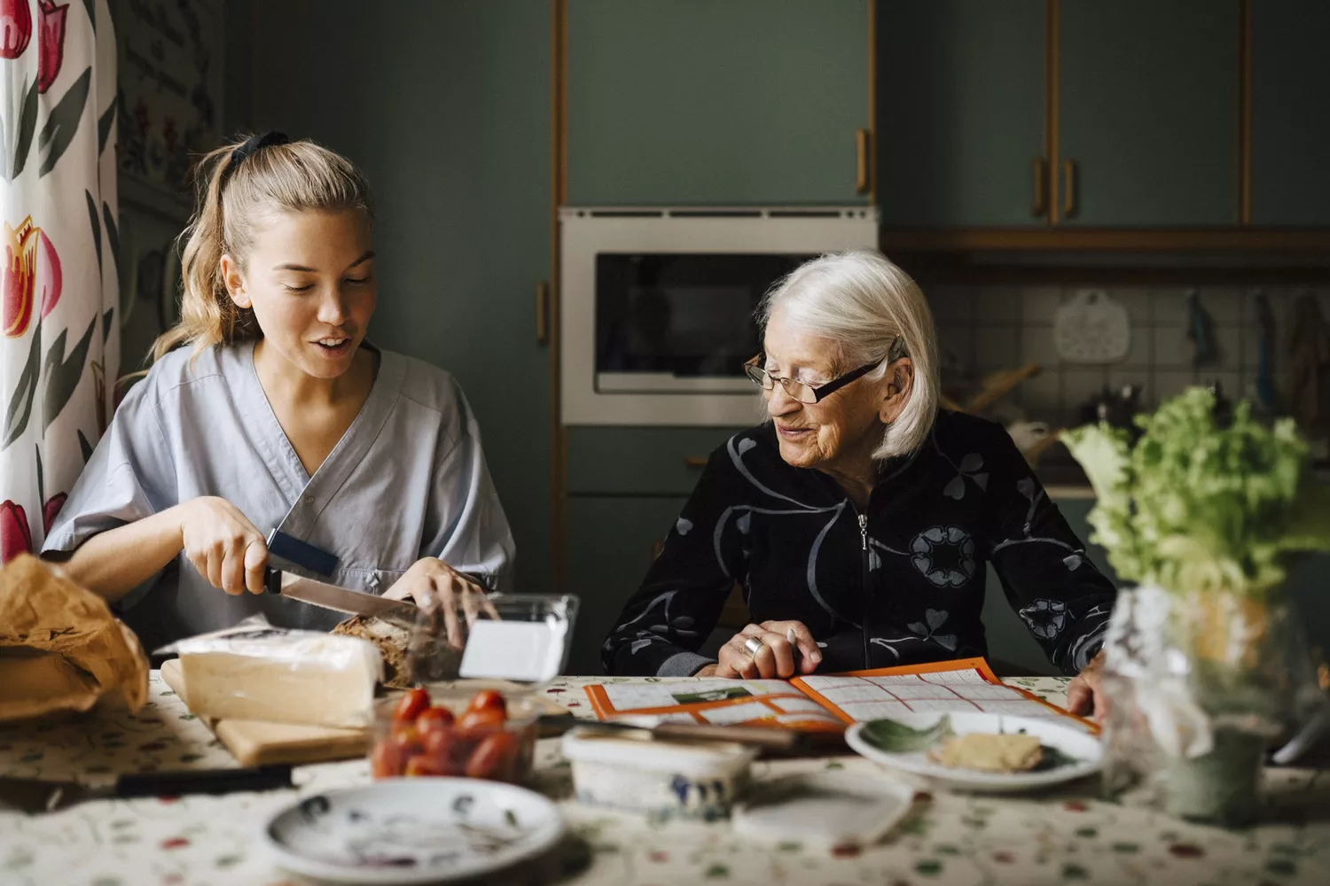 A female caregiver cutting bread for a senior woman in a well-lit kitchen.