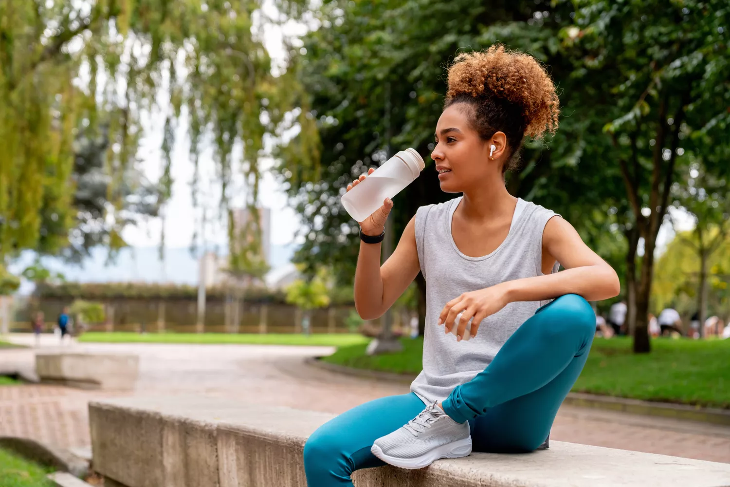 A person in athletic wear drinking water from a clear bottle while outdoors in a park.