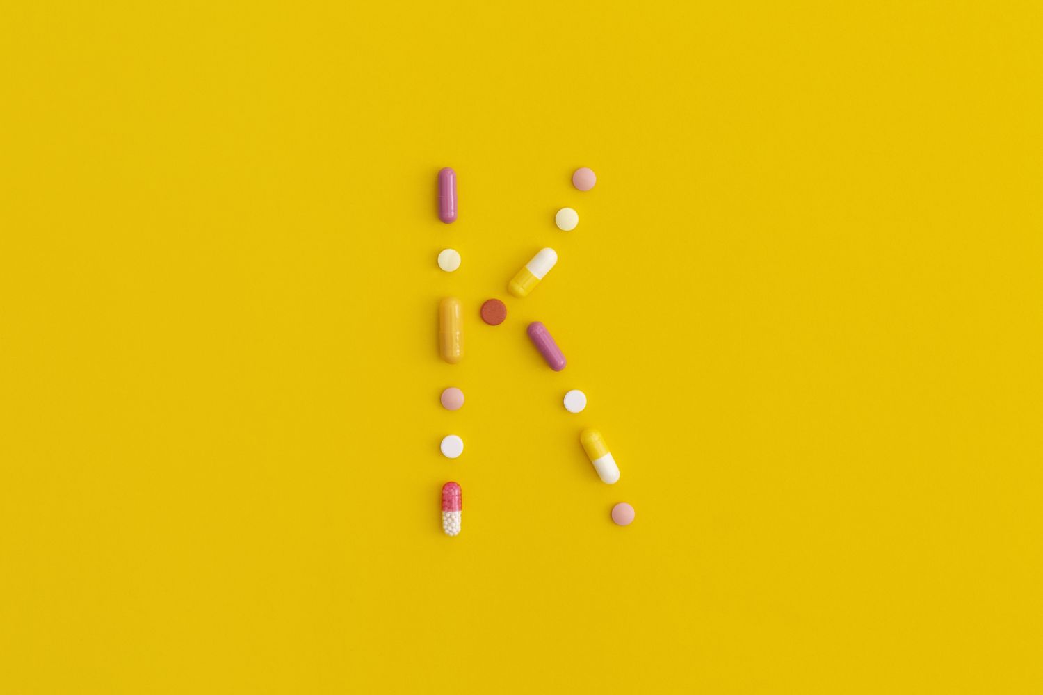 Vitamin K2 pills and tablets arranged in the shape of the letter K on a bright yellow surface.