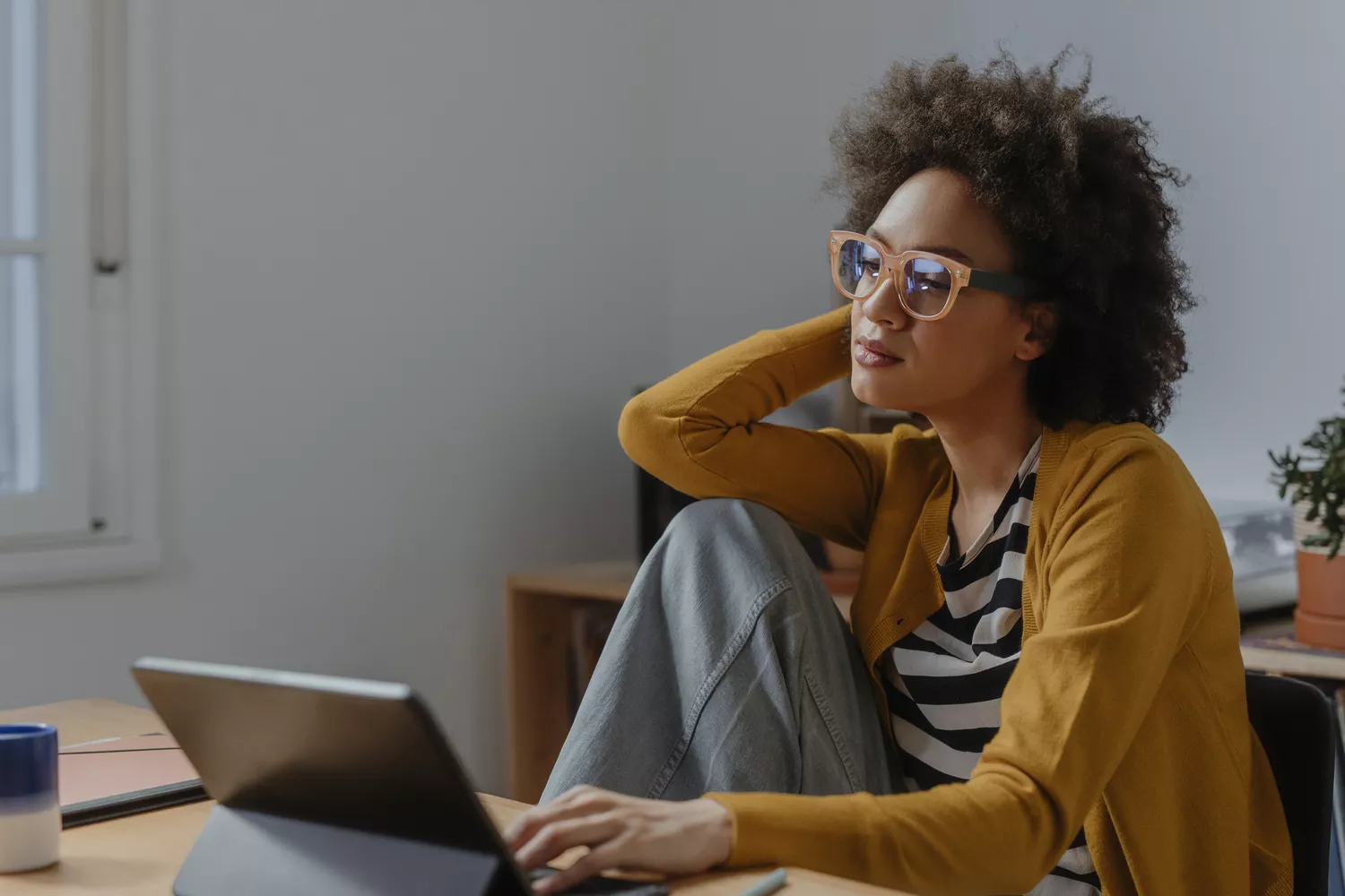 A woman wearing glasses resting her head on her hand while looking thoughtfully at her laptop screen.